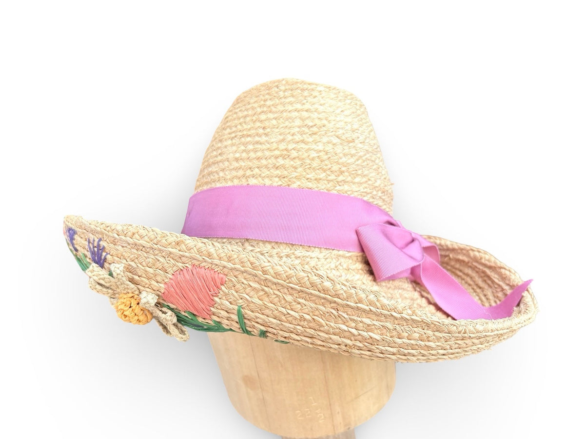 A straw sun hat with a wide brim, decorated with a pink ribbon and bow, and embroidered with colorful flowers on the side, displayed on a wooden mannequin head against a white background.