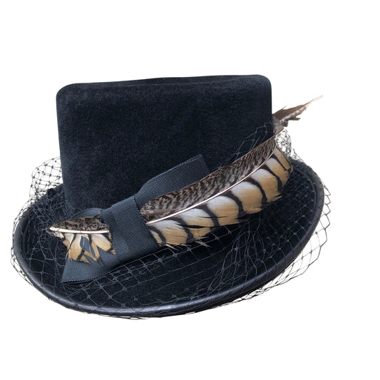 A Victorian-inspired top hat, made-to-order "Pickwick" hat from Cha Cha's House of Ill Repute, a woman-owned millinery in New York City. The coachman-style hat is crafted in black with a slightly lower profile than a traditional top hat. It's adorned with an elegant, dotted black veiling and features a decorative pheasant tail feather, showcasing natural brown and cream patterns. The hat is accented with a black ribbon band, creating a piece that blends historical charm with contemporary fashion.