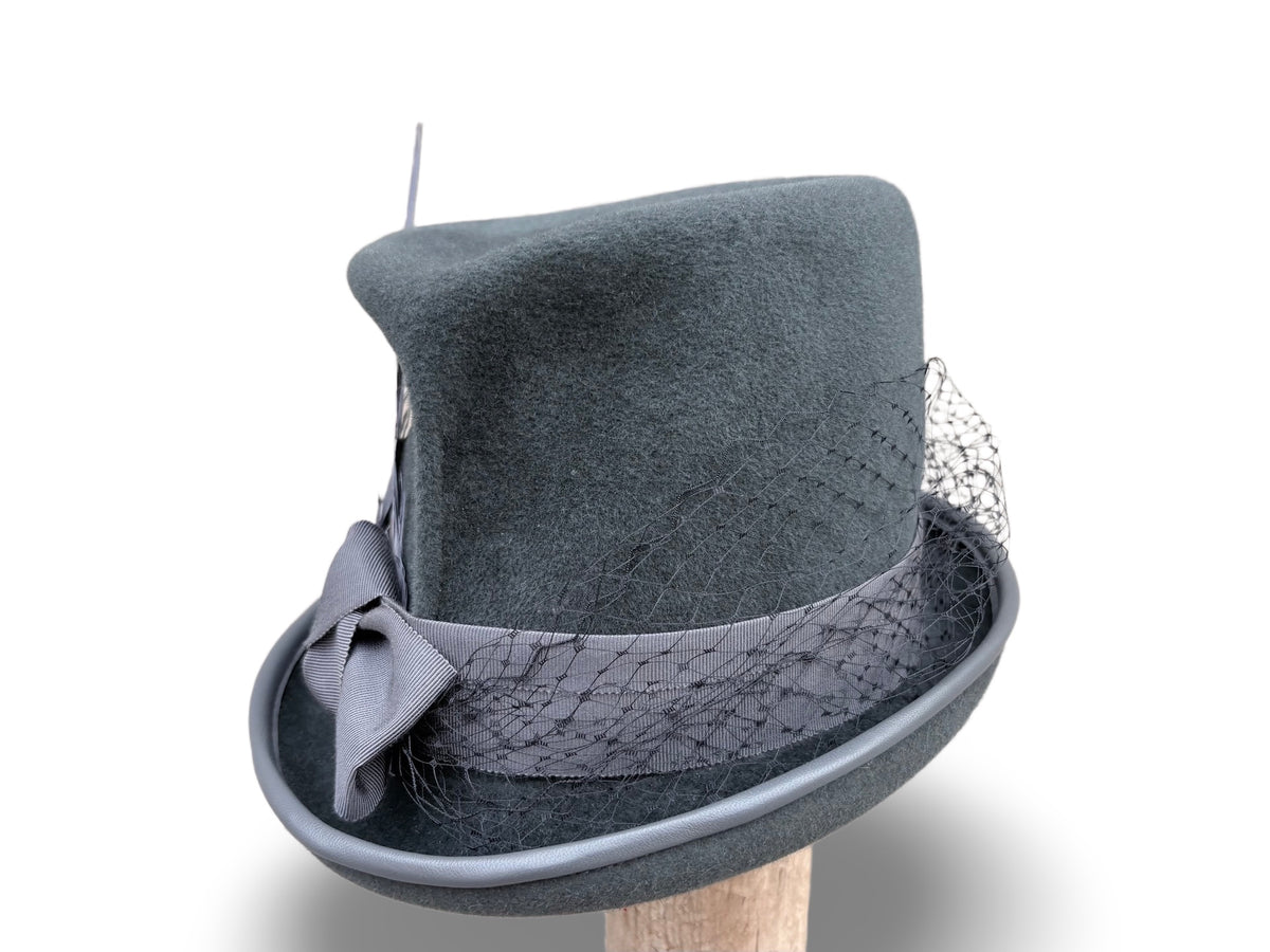 A dark gray felt top hat with a matching gray ribbon, bow, and delicate black mesh veil, displayed on a wooden stand against a white background.