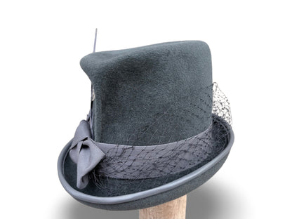 A dark gray felt top hat with a matching gray ribbon, bow, and delicate black mesh veil, displayed on a wooden stand against a white background.