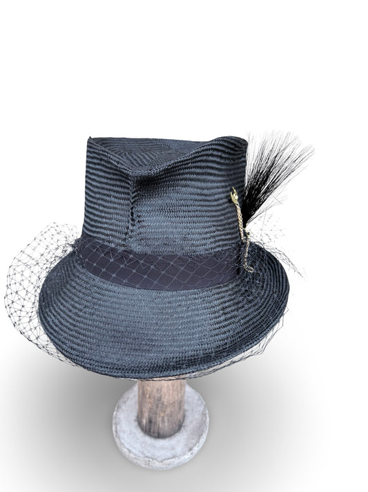 A black parisisol straw fedora hat with a black, black feather accent, and white netting veil, displayed on a wooden stand against a white background