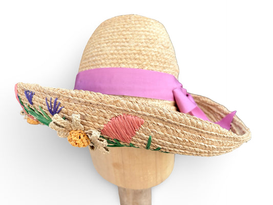 A woven straw sun hat displayed on a wooden mannequin head against a white background. The hat features a vibrant pink ribbon band and a wide brim decorated with colorful floral embroidery in pink, yellow, purple, and green.