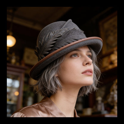 A woman with short gray hair wearing a dark gray felt Cloche hat with a brown leather piping and leaf embellishments, photographed in a warm, vintage-style shop interior.