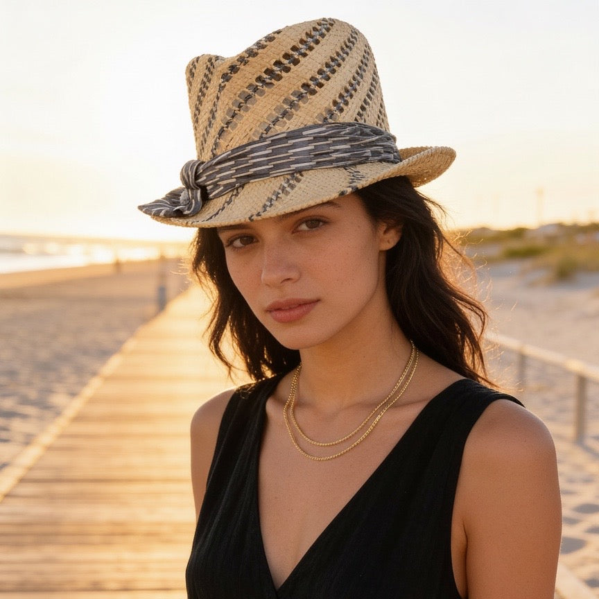 A woman wearing a woven straw fedora hat with a patterned band and a black sleeveless dress, standing on a wooden boardwalk at sunset.