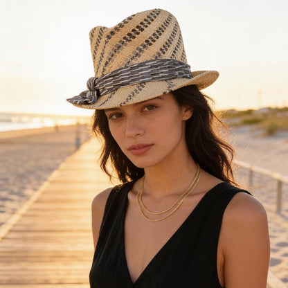A woman wearing a woven straw fedora hat with a patterned band and a black sleeveless dress, standing on a wooden boardwalk at sunset.