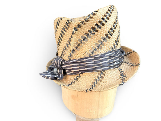 A woven straw fedora hat with black and white striped pattern details and a matching patterned band, displayed on a wooden stand against a white background.