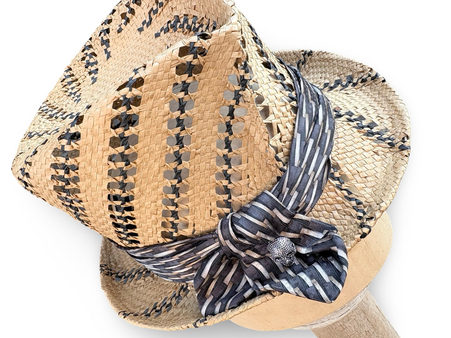 A woven straw fedora hat with black and beige geometric patterns, featuring a navy and beige striped silk ribbon tied in a bow with a silver decorative center.