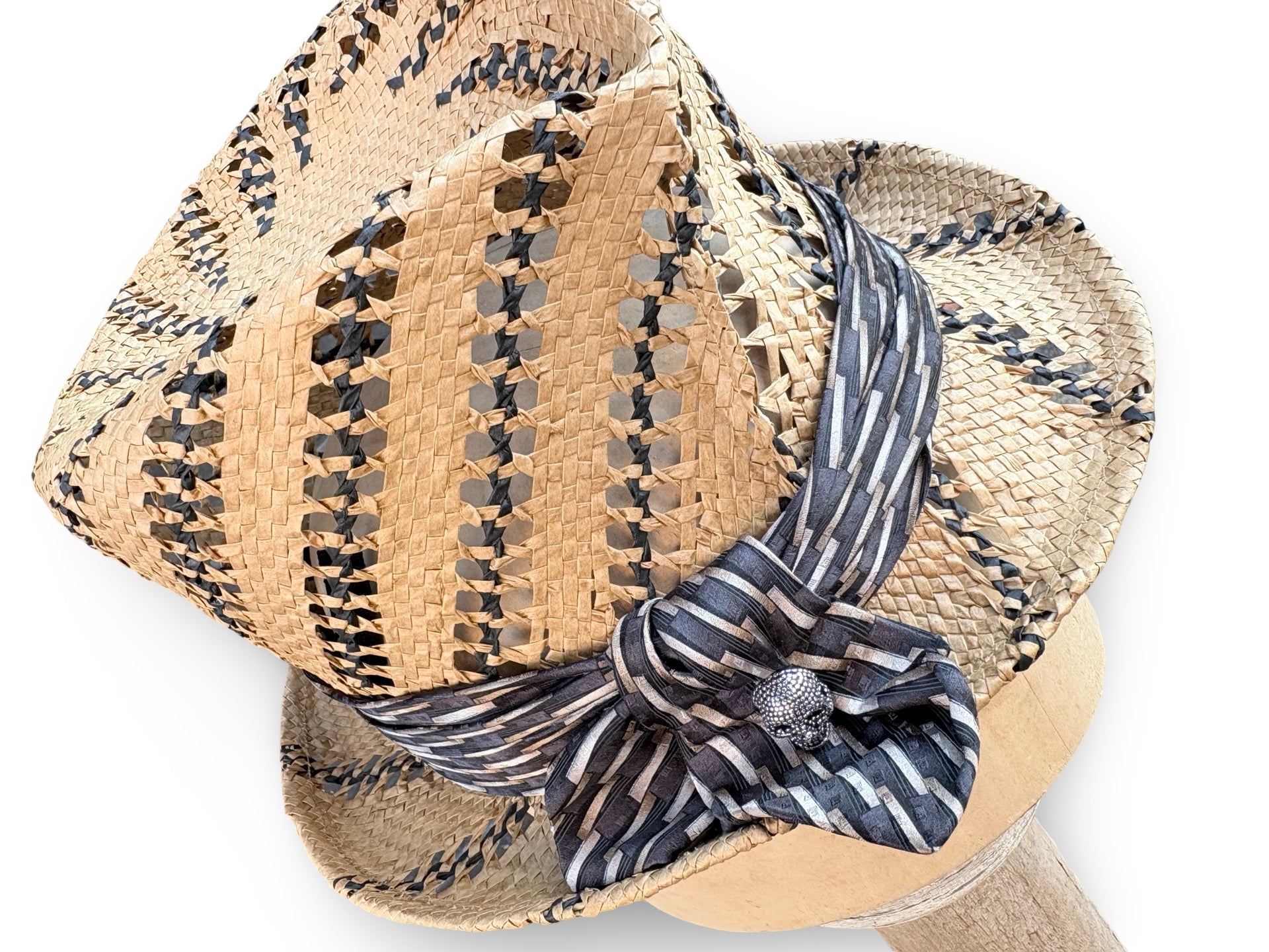 A woven straw fedora hat with black and beige geometric patterns, featuring a navy and beige striped silk ribbon tied in a bow with a silver decorative center.
