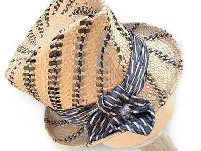 A woven straw fedora hat with black and beige geometric patterns, featuring a navy and beige striped silk ribbon tied in a bow with a silver decorative center.