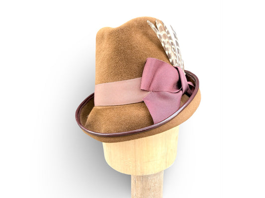 A bronze felt hat with a dusty tonal ribbon band, a matching rust bow, and a feather accent, displayed on a wooden stand against a white background.