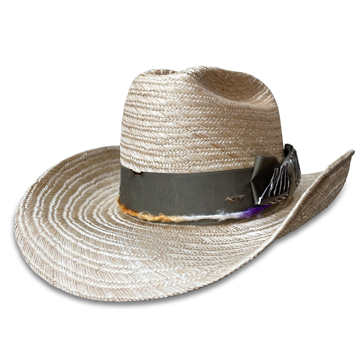 A close-up of a straw cowboy hat, La Chatte, featuring a distressed grosgrain ribbon and colorful, hand-dyed silk yarn accents. A turkey wing feather adds a unique touch to the side of the hat.