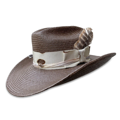 Dark brown straw cowboy hat with a distressed look, ribbon and silk embellishments, and a feather accent